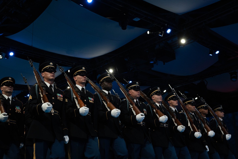 A military honor guard unit stands in a precise formation on a stage under dramatic blue and white stage lighting. The soldiers are dressed in formal dark dress uniforms with service medals, white gloves, and black service caps, each holding a rifle at the same angle. The setting appears to be an indoor ceremonial event or formal military presentation.