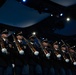 A military honor guard unit stands in a precise formation on a stage under dramatic blue and white stage lighting. The soldiers are dressed in formal dark dress uniforms with service medals, white gloves, and black service caps, each holding a rifle at the same angle. The setting appears to be an indoor ceremonial event or formal military presentation.