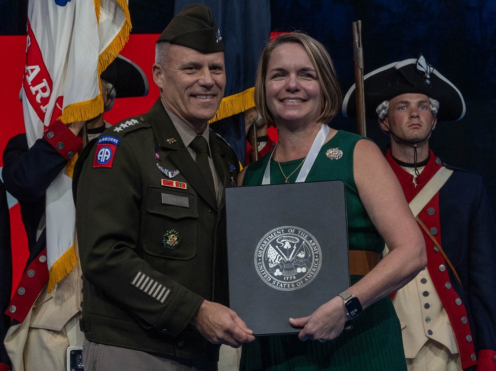 A U.S. Army general officer and a civilian woman pose together holding a Department of the Army certificate folder during what appears to be a formal military ceremony or awards presentation. The general is wearing an Army Service Uniform with an 82nd Airborne Division patch and four-star rank insignia. In the background, a soldier dressed in a Revolutionary War-era uniform stands at attention alongside military color guard flags.
