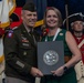 A U.S. Army general officer and a civilian woman pose together holding a Department of the Army certificate folder during what appears to be a formal military ceremony or awards presentation. The general is wearing an Army Service Uniform with an 82nd Airborne Division patch and four-star rank insignia. In the background, a soldier dressed in a Revolutionary War-era uniform stands at attention alongside military color guard flags.