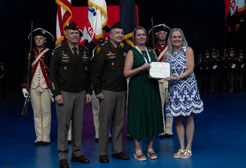 A formal military awards ceremony is taking place on a stage with a blue floor. Two Army officers in dress uniforms are posing with two civilian women who are jointly holding a certificate or award. The backdrop features multiple military flags and a color guard in both modern Army uniforms and historical Revolutionary War-era uniforms.