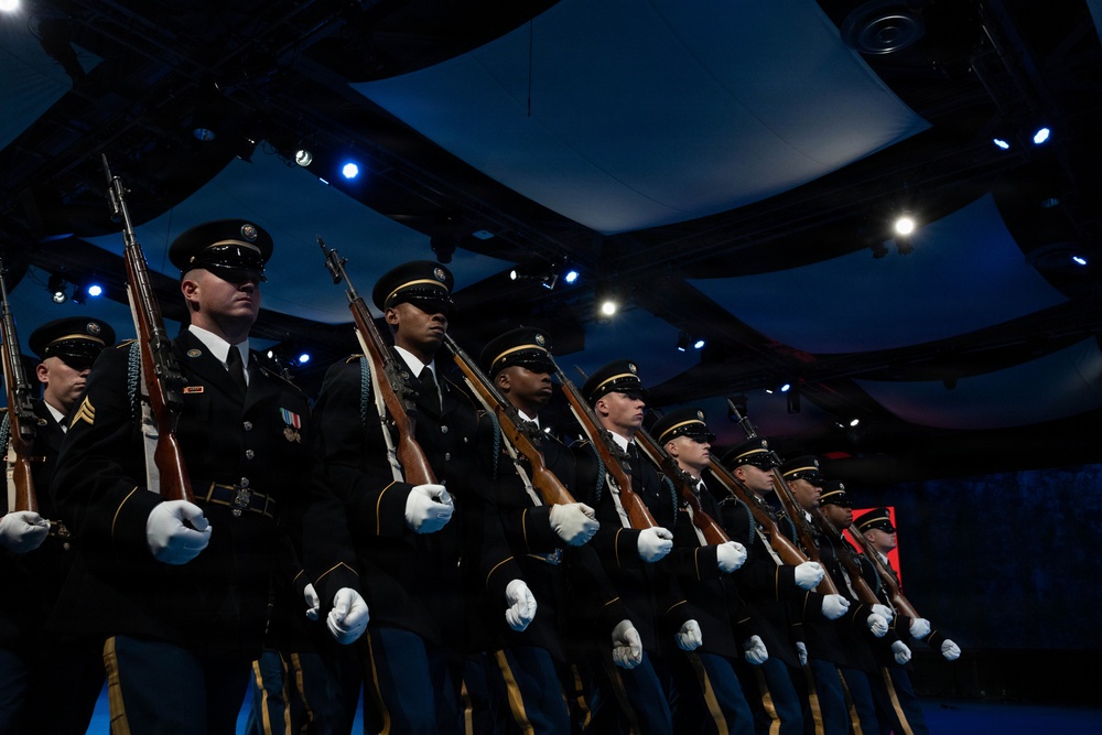 A military honor guard unit stands in a precise diagonal formation inside an indoor venue with dramatic stage lighting. The soldiers are dressed in formal dark dress uniforms, wearing white gloves, and carrying rifles at a uniform angle. The setting appears to be a formal ceremonial event or performance.