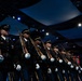 A military honor guard unit stands in a precise diagonal formation inside an indoor venue with dramatic stage lighting. The soldiers are dressed in formal dark dress uniforms, wearing white gloves, and carrying rifles at a uniform angle. The setting appears to be a formal ceremonial event or performance.