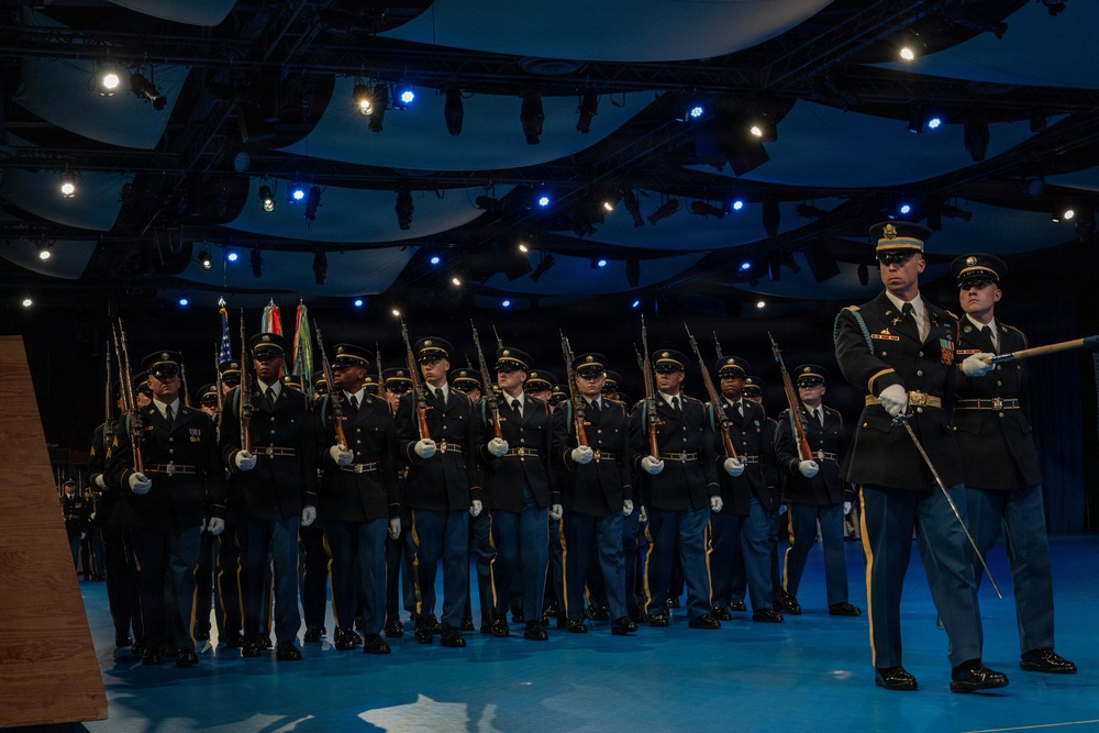 A formal military ceremony is taking place on an indoor stage with dramatic blue lighting. A large formation of uniformed soldiers stands at attention holding rifles, with color guard members carrying flags visible in the background. Two officers in dress uniforms stand at the front right, one holding a ceremonial sword or baton.