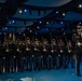 A formal military ceremony is taking place on an indoor stage with dramatic blue lighting. A large formation of uniformed soldiers stands at attention holding rifles, with color guard members carrying flags visible in the background. Two officers in dress uniforms stand at the front right, one holding a ceremonial sword or baton.