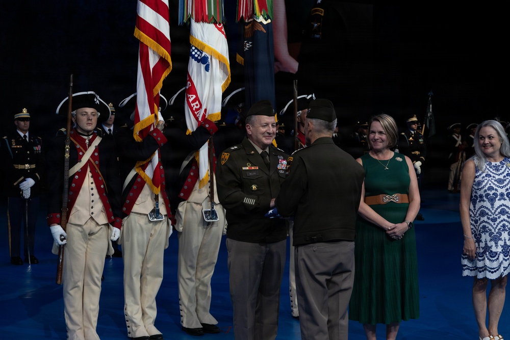 A formal U.S. Army military ceremony is taking place on a stage with a blue floor. Two officers in modern Army Combat Uniforms are exchanging a folded American flag, flanked by color guard soldiers in historical Revolutionary War-era uniforms. Two civilian women in formal attire stand nearby as witnesses to the ceremony.