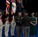 A formal U.S. Army military ceremony is taking place on a stage with a blue floor. Two officers in modern Army Combat Uniforms are exchanging a folded American flag, flanked by color guard soldiers in historical Revolutionary War-era uniforms. Two civilian women in formal attire stand nearby as witnesses to the ceremony.