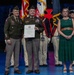 A formal U.S. Army retirement ceremony is taking place on a stage. A uniformed officer in the center is holding a Certificate of Retirement, flanked by a senior officer on the left and a civilian woman in a green dress on the right. A military honor guard in historical uniforms stands in the background alongside U.S. and unit flags.