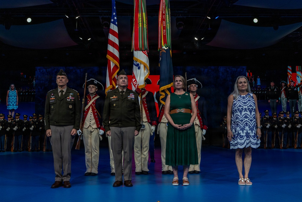 A formal military ceremony is taking place on a stage with blue lighting. Four principal figures — two Army officers in dress uniforms and two civilian women — stand at attention in the foreground, flanked by a color guard in Revolutionary War-era uniforms holding the U.S. flag and military unit colors. Rows of uniformed soldiers are visible in the background, suggesting a large-scale official event such as a change of command or military anniversary ceremony.