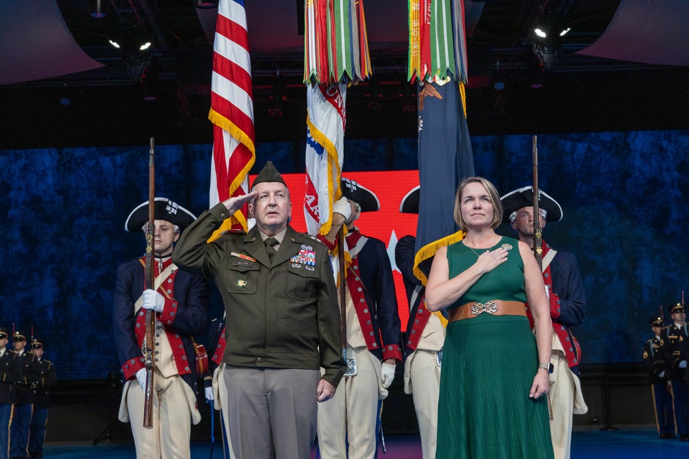 A formal military ceremony is taking place on a stage, featuring a senior military officer in dress uniform rendering a salute alongside a civilian woman in a green dress with her hand over her heart. Behind them stands a color guard in historical Continental Army uniforms holding multiple flags including the American flag. The setting appears to be a large indoor venue with dramatic lighting.