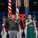 A formal military ceremony is taking place on a stage, featuring a senior military officer in dress uniform rendering a salute alongside a civilian woman in a green dress with her hand over her heart. Behind them stands a color guard in historical Continental Army uniforms holding multiple flags including the American flag. The setting appears to be a large indoor venue with dramatic lighting.