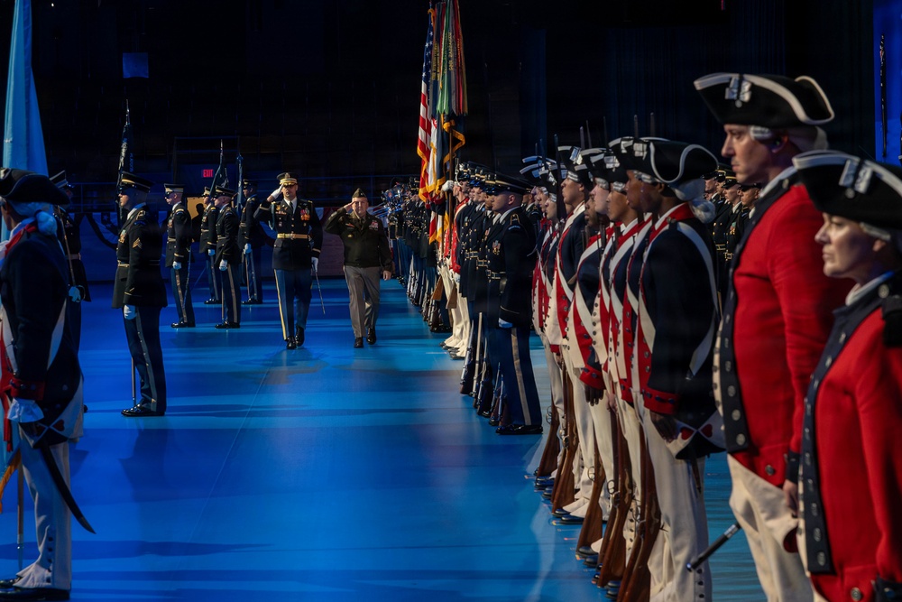 A formal military ceremony is taking place in a large indoor venue with blue-lit flooring. Two senior officers are walking down a central aisle being saluted, flanked by rows of soldiers in both modern dress uniforms and historical Revolutionary War-era uniforms. The event appears to be a high-level military review or change of command ceremony.
