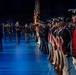 A formal military ceremony is taking place in a large indoor venue with blue-lit flooring. Two senior officers are walking down a central aisle being saluted, flanked by rows of soldiers in both modern dress uniforms and historical Revolutionary War-era uniforms. The event appears to be a high-level military review or change of command ceremony.