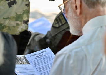An elderly man is reading a printed program or pamphlet titled 'Remembering The Battle of Bladensburg' at what appears to be a commemorative event. A person in military camouflage uniform is visible in the background, suggesting this is an official military or historical ceremony. The scene depicts attendees gathered for a memorial or anniversary observance related to the War of 1812 Battle of Bladensburg.