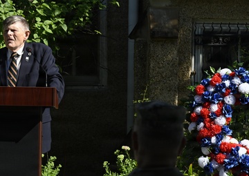 An elderly man in a dark suit and striped tie delivers a speech at a wooden podium during what appears to be a memorial ceremony. A large red, white, and blue floral wreath is prominently displayed to the right, consistent with American patriotic or military commemorative events. Military personnel in camouflage uniforms are visible in the foreground as attendees.