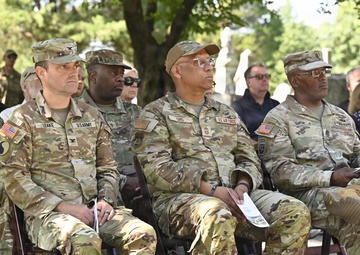 A group of U.S. military personnel from multiple branches, including Army and Air Force, are seated in folding chairs at an outdoor ceremony, likely a memorial or commemorative event. Several name tapes are visible including LEAKE, SIMS, and SMITH. Civilians are also present in the background, and the setting appears to be a cemetery or memorial grounds.