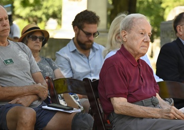 A group of people are seated in wooden folding chairs outdoors, appearing to attend a formal ceremony or memorial event. The setting appears to be a cemetery or memorial garden, visible from the stone markers in the background. Attendees range in age and are dressed in casual to semi-formal attire, with most appearing attentive.