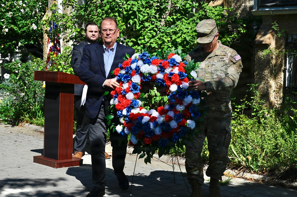 A memorial or commemorative ceremony is taking place outdoors, featuring a civilian in a dark suit and a U.S. Army soldier in camouflage uniform jointly handling a red, white, and blue floral wreath. A podium is visible to the left, and a third individual stands in the background. The wreath's patriotic colors suggest this is a military or national remembrance event.