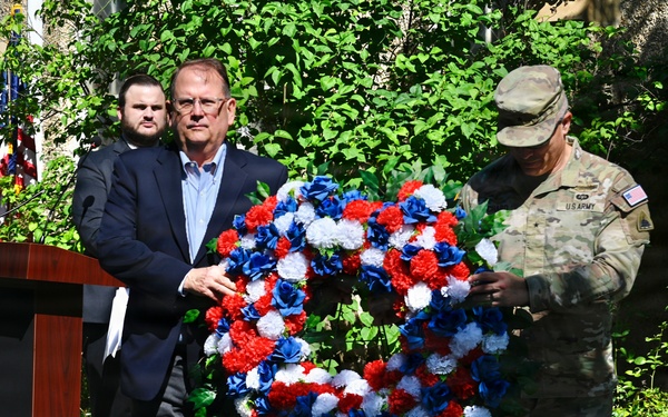 A memorial or commemorative ceremony is taking place outdoors, featuring a civilian in a dark suit and a U.S. Army soldier in camouflage uniform jointly handling a red, white, and blue floral wreath. A podium is visible to the left, and a third individual stands in the background. The wreath's patriotic colors suggest this is a military or national remembrance event.