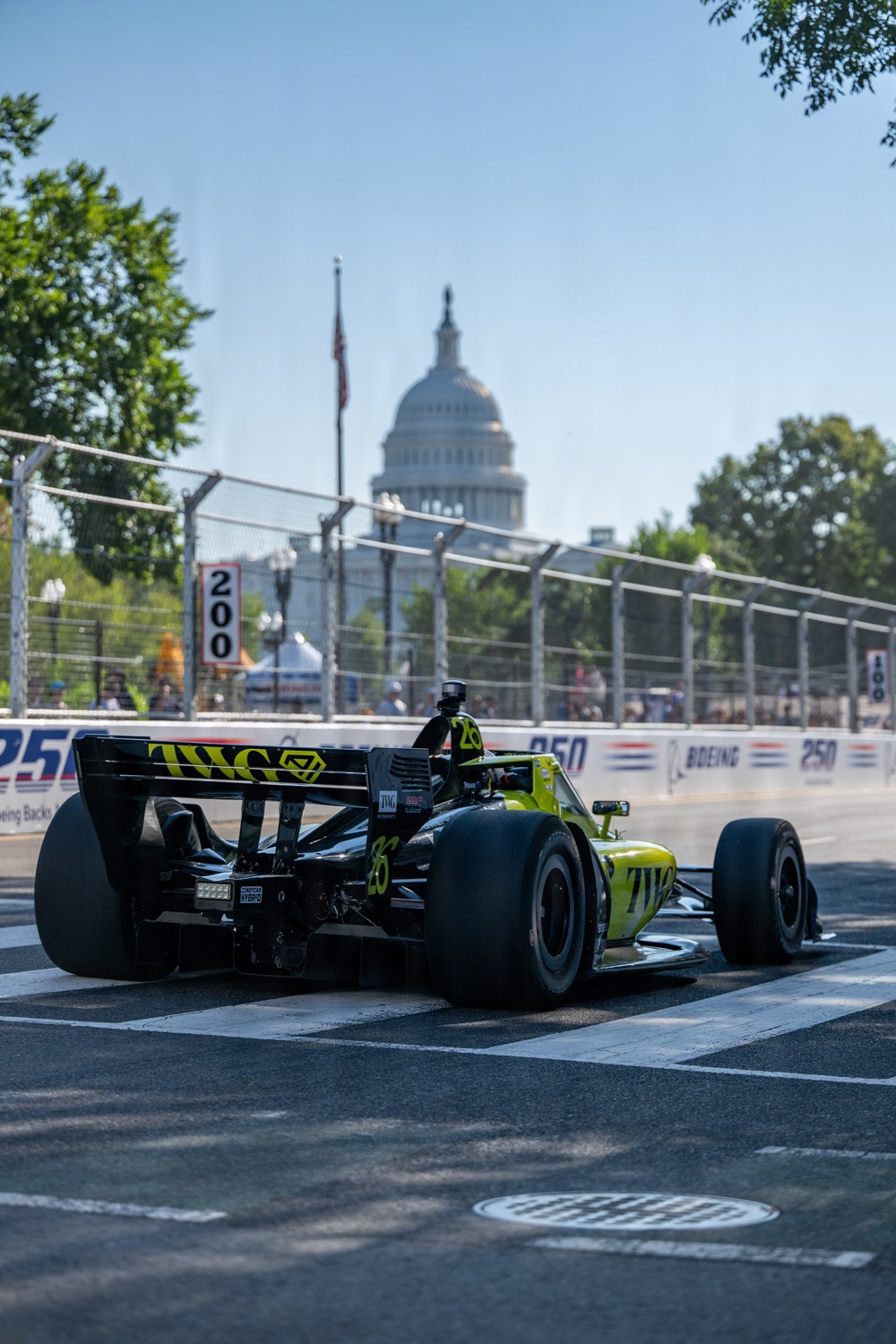An IndyCar racing car with yellow-green and black livery bearing the 'TWG' sponsorship and number 26 is navigating a street circuit. The race is taking place in Washington D.C., with the United States Capitol building clearly visible in the background. Sponsor banners reading '250' and 'Boeing' line the temporary circuit barriers.