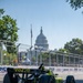 An IndyCar racing car with yellow-green and black livery bearing the 'TWG' sponsorship and number 26 is navigating a street circuit. The race is taking place in Washington D.C., with the United States Capitol building clearly visible in the background. Sponsor banners reading '250' and 'Boeing' line the temporary circuit barriers.