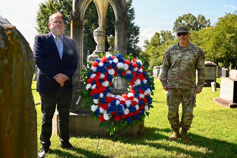 Two individuals stand beside a red, white, and blue floral wreath at a cemetery monument, suggesting a memorial or commemorative ceremony. One person is in civilian attire wearing a dark suit, while the other is in U.S. Army combat uniform with a name tape reading 'BLANCHARD'. The setting includes historic gravestones and a Gothic-style stone monument in the background.