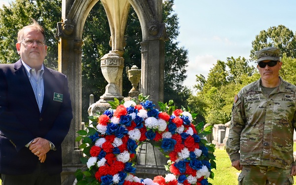 Two individuals stand beside a red, white, and blue floral wreath at a cemetery monument, suggesting a memorial or commemorative ceremony. One person is in civilian attire wearing a dark suit, while the other is in U.S. Army combat uniform with a name tape reading 'BLANCHARD'. The setting includes historic gravestones and a Gothic-style stone monument in the background.