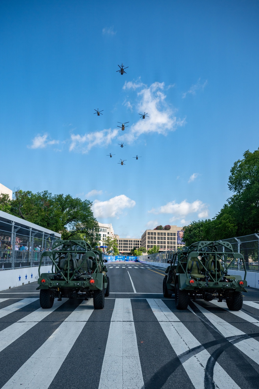 A military display at what appears to be a motorsport racing event, featuring two olive-drab military light tactical vehicles (likely Ground Mobility Vehicles or similar) positioned on a race track starting grid. A formation of approximately 9-10 military helicopters flies overhead in a diagonal echelon formation against a blue sky. Sponsor banners reading 'NTT' are visible in the background, suggesting this is an IndyCar or similar racing series event.