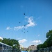 A military display at what appears to be a motorsport racing event, featuring two olive-drab military light tactical vehicles (likely Ground Mobility Vehicles or similar) positioned on a race track starting grid. A formation of approximately 9-10 military helicopters flies overhead in a diagonal echelon formation against a blue sky. Sponsor banners reading 'NTT' are visible in the background, suggesting this is an IndyCar or similar racing series event.