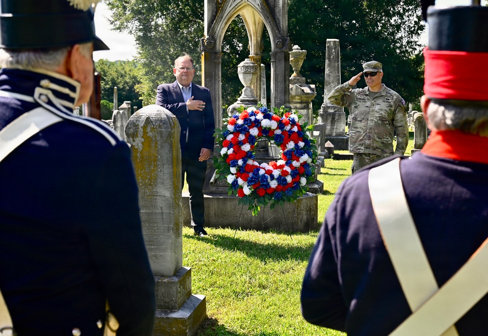 A memorial ceremony is taking place in a historic cemetery, featuring a red, white, and blue floral wreath placed against a gravestone. A civilian in a suit has his hand over his heart while a military officer in modern camouflage uniform renders a salute. Two individuals in historical military reenactor uniforms (dark blue coats with red shakos) are visible in the foreground.