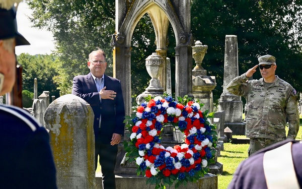 A memorial ceremony is taking place in a historic cemetery, featuring a red, white, and blue floral wreath placed against a gravestone. A civilian in a suit has his hand over his heart while a military officer in modern camouflage uniform renders a salute. Two individuals in historical military reenactor uniforms (dark blue coats with red shakos) are visible in the foreground.