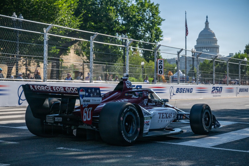 An IndyCar race car numbered 60, sponsored by Texas A&amp;M University and bearing 'A Force for Good' livery, navigates a street circuit in Washington D.C. The U.S. Capitol building is clearly visible in the background, identifying this as the former IndyCar Grand Prix on the Streets of Washington D.C. Trackside barriers display Boeing sponsorship branding alongside distance markers.