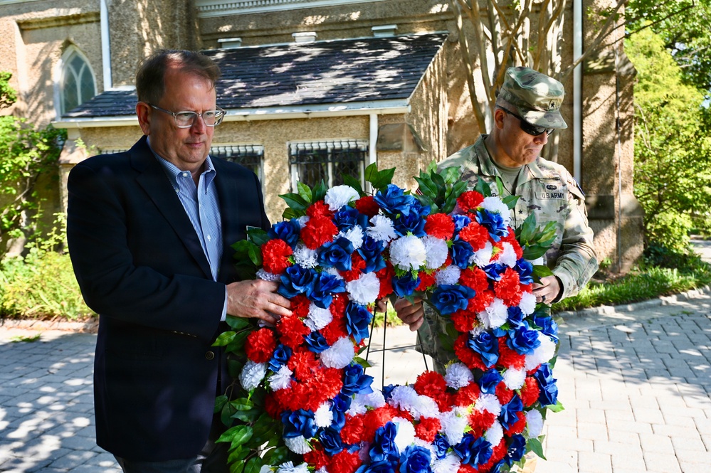 Two individuals are carrying a large patriotic wreath composed of red, white, and blue flowers during what appears to be a memorial or commemorative ceremony. One person is in civilian attire (dark blazer, blue shirt, glasses) and the other is wearing a U.S. Army camouflage uniform with rank insignia. The setting is outdoors near a stone building that resembles a church or historic structure.