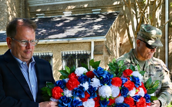 Two individuals are carrying a large patriotic wreath composed of red, white, and blue flowers during what appears to be a memorial or commemorative ceremony. One person is in civilian attire (dark blazer, blue shirt, glasses) and the other is wearing a U.S. Army camouflage uniform with rank insignia. The setting is outdoors near a stone building that resembles a church or historic structure.