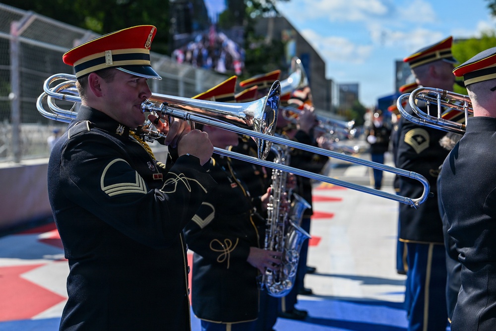 A military marching band is performing outdoors, likely at a public event or ceremony. The musicians are dressed in formal dark uniforms with distinctive red and black caps featuring gold trim. The setting appears to be near a racing circuit or urban venue, based on the track markings and barriers visible in the background.