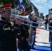 A military marching band is performing outdoors, likely at a public event or ceremony. The musicians are dressed in formal dark uniforms with distinctive red and black caps featuring gold trim. The setting appears to be near a racing circuit or urban venue, based on the track markings and barriers visible in the background.