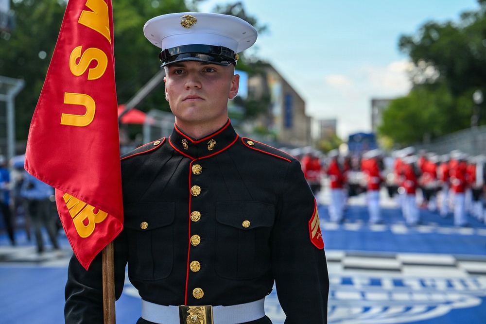 A United States Marine in dress blues uniform stands at attention holding a red guidon flag with visible letters 'U' and 'MB'. In the background, a formation of Marines in matching dress uniforms with red jackets and white trousers are assembled on a blue surface, suggesting a formal ceremony or parade. The setting appears to be an outdoor urban environment with trees and buildings visible.