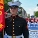 A United States Marine in dress blues uniform stands at attention holding a red guidon flag with visible letters 'U' and 'MB'. In the background, a formation of Marines in matching dress uniforms with red jackets and white trousers are assembled on a blue surface, suggesting a formal ceremony or parade. The setting appears to be an outdoor urban environment with trees and buildings visible.