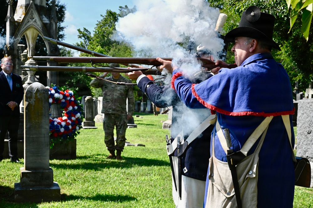 A ceremonial musket volley is being fired at a cemetery by individuals dressed in early American (Revolutionary War-era) military reenactment uniforms. The event appears to be a memorial or commemorative ceremony, evidenced by a red, white, and blue wreath placed near a gravestone. A modern military service member in camouflage uniform and civilians are visible observing in the background.