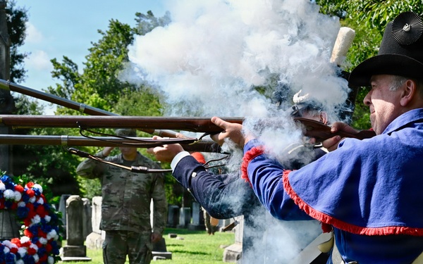 A ceremonial musket volley is being fired at a cemetery by individuals dressed in early American (Revolutionary War-era) military reenactment uniforms. The event appears to be a memorial or commemorative ceremony, evidenced by a red, white, and blue wreath placed near a gravestone. A modern military service member in camouflage uniform and civilians are visible observing in the background.