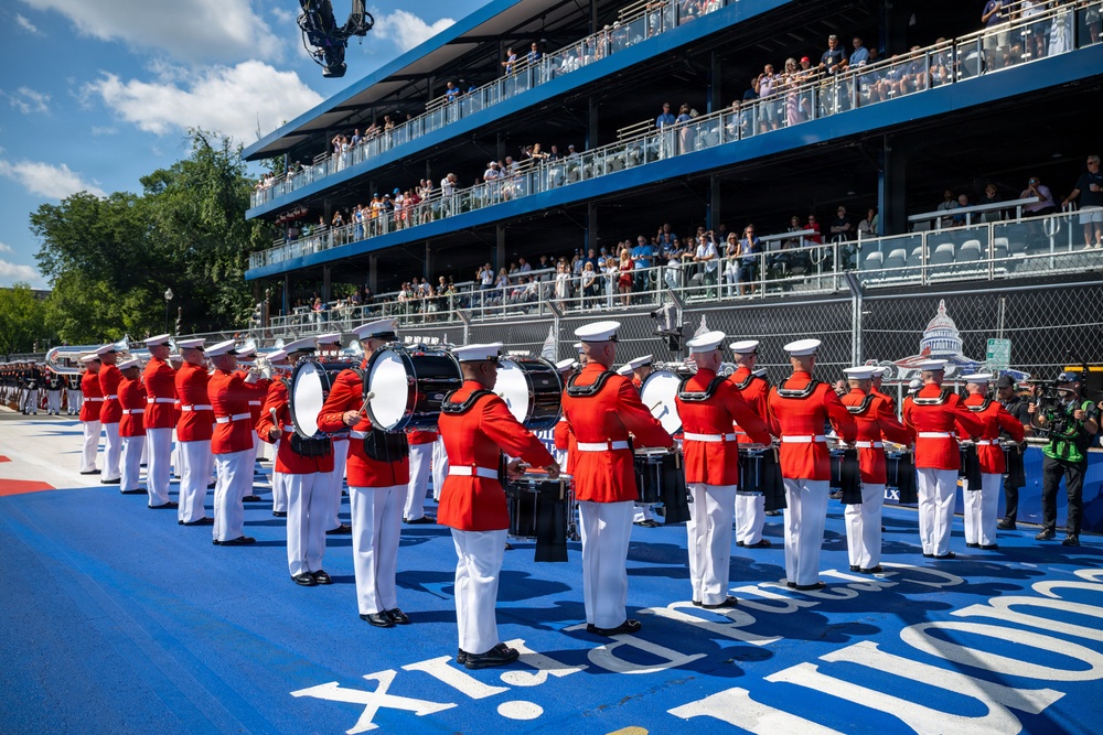 A military marching band, identifiable by their distinctive red tunics and white trousers with white peaked caps, performs on a blue surface at what appears to be a large outdoor event venue. The performers are arranged in formation with various instruments including bass drums and brass instruments. A multi-tiered grandstand filled with spectators is visible in the background, along with what appears to be the U.S. Capitol dome in the far right background.