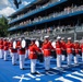 A military marching band, identifiable by their distinctive red tunics and white trousers with white peaked caps, performs on a blue surface at what appears to be a large outdoor event venue. The performers are arranged in formation with various instruments including bass drums and brass instruments. A multi-tiered grandstand filled with spectators is visible in the background, along with what appears to be the U.S. Capitol dome in the far right background.