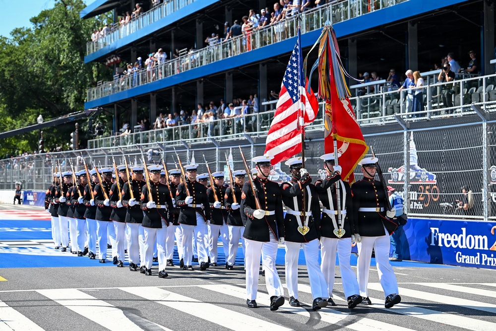 A United States Marine Corps Silent Drill Platoon or Honor Guard is marching in ceremonial formation at what appears to be the 'Freedom Grand Prix' racing event. The Marines are dressed in full dress blues with white trousers, carrying rifles and color guard flags including the American flag and a Marine Corps flag. Spectators fill a multi-tiered grandstand in the background.