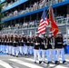 A United States Marine Corps Silent Drill Platoon or Honor Guard is marching in ceremonial formation at what appears to be the 'Freedom Grand Prix' racing event. The Marines are dressed in full dress blues with white trousers, carrying rifles and color guard flags including the American flag and a Marine Corps flag. Spectators fill a multi-tiered grandstand in the background.