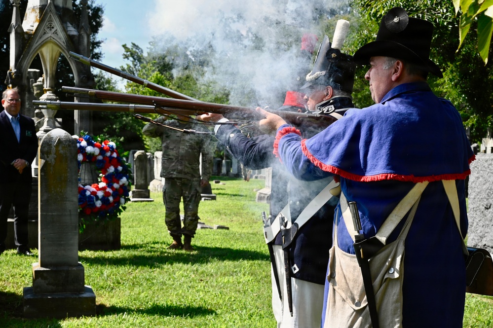 A military memorial ceremony is taking place in a historic cemetery, where reenactors dressed in early American (Revolutionary War-era) uniforms are firing muskets in a ceremonial volley salute. A patriotic red, white, and blue wreath is displayed near a gravestone, and a modern military service member in camouflage uniform stands in the background alongside a civilian. The smoke from the discharged muskets is clearly visible, indicating the weapons have just been fired.