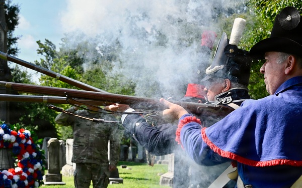 A military memorial ceremony is taking place in a historic cemetery, where reenactors dressed in early American (Revolutionary War-era) uniforms are firing muskets in a ceremonial volley salute. A patriotic red, white, and blue wreath is displayed near a gravestone, and a modern military service member in camouflage uniform stands in the background alongside a civilian. The smoke from the discharged muskets is clearly visible, indicating the weapons have just been fired.