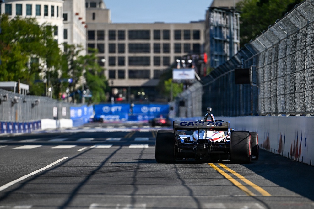 An IndyCar racing event taking place on a temporary street circuit in an urban environment. The foreground features a single-seater race car with 'GAYLOR' sponsorship livery and patriotic red, white, and blue coloring, viewed from the rear. The track is lined with concrete barriers and catch fencing, with city buildings visible in the background.