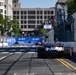 An IndyCar racing event taking place on a temporary street circuit in an urban environment. The foreground features a single-seater race car with 'GAYLOR' sponsorship livery and patriotic red, white, and blue coloring, viewed from the rear. The track is lined with concrete barriers and catch fencing, with city buildings visible in the background.