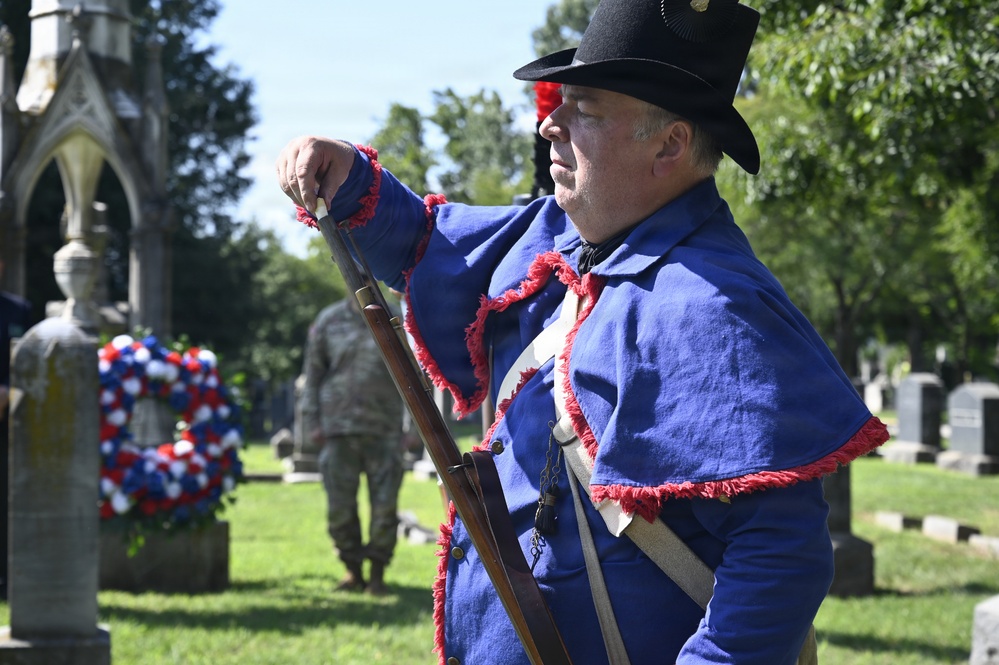 A historical reenactor dressed in a blue uniform with red fringe trim and a black hat stands at attention holding a period musket in a cemetery setting. In the background, a red, white, and blue wreath is placed near a stone monument, and a modern military soldier in camouflage uniform is visible. The scene appears to be a memorial or commemorative ceremony honoring military veterans.
