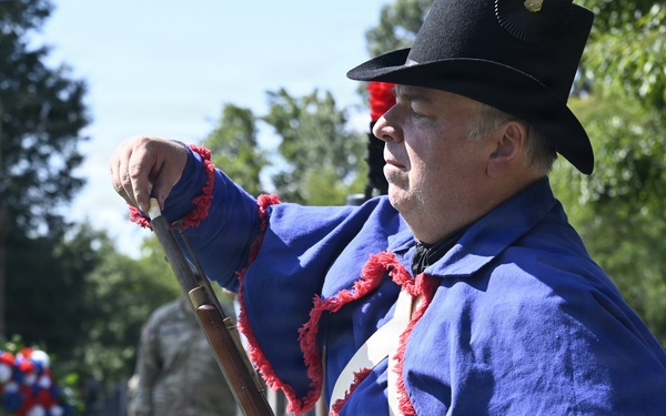 A historical reenactor dressed in a blue uniform with red fringe trim and a black hat stands at attention holding a period musket in a cemetery setting. In the background, a red, white, and blue wreath is placed near a stone monument, and a modern military soldier in camouflage uniform is visible. The scene appears to be a memorial or commemorative ceremony honoring military veterans.